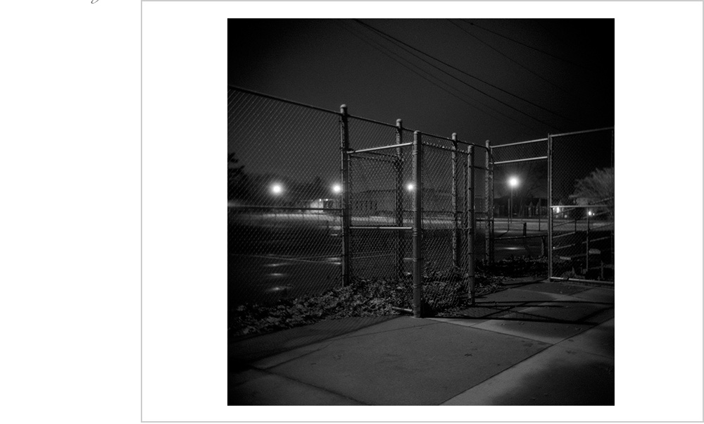 The solitude of a tennis court at night with only power lines and chainlink fence to occupy.